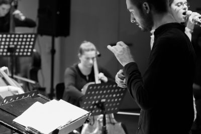 Dirigent vor Pult, Ensemble und S&auml;ngerin im Hintergrund | Copyright: Hennri Kleemann