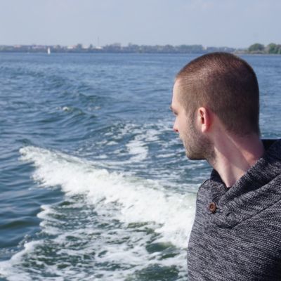 Marius Moritz blickt von einem Boot aus auf das Wasser, im Hintergrund ein Segelboot und die Uferlinie unter blauem Himmel. | Copyright: Marius Moritz