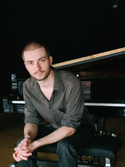 Marius Moritz sitzt in dunklem Hemd auf einem Hocker vor einem Fl&uuml;gel, H&auml;nde gefaltet, Blick in die Kamera. | Copyright: Thomas Sasse